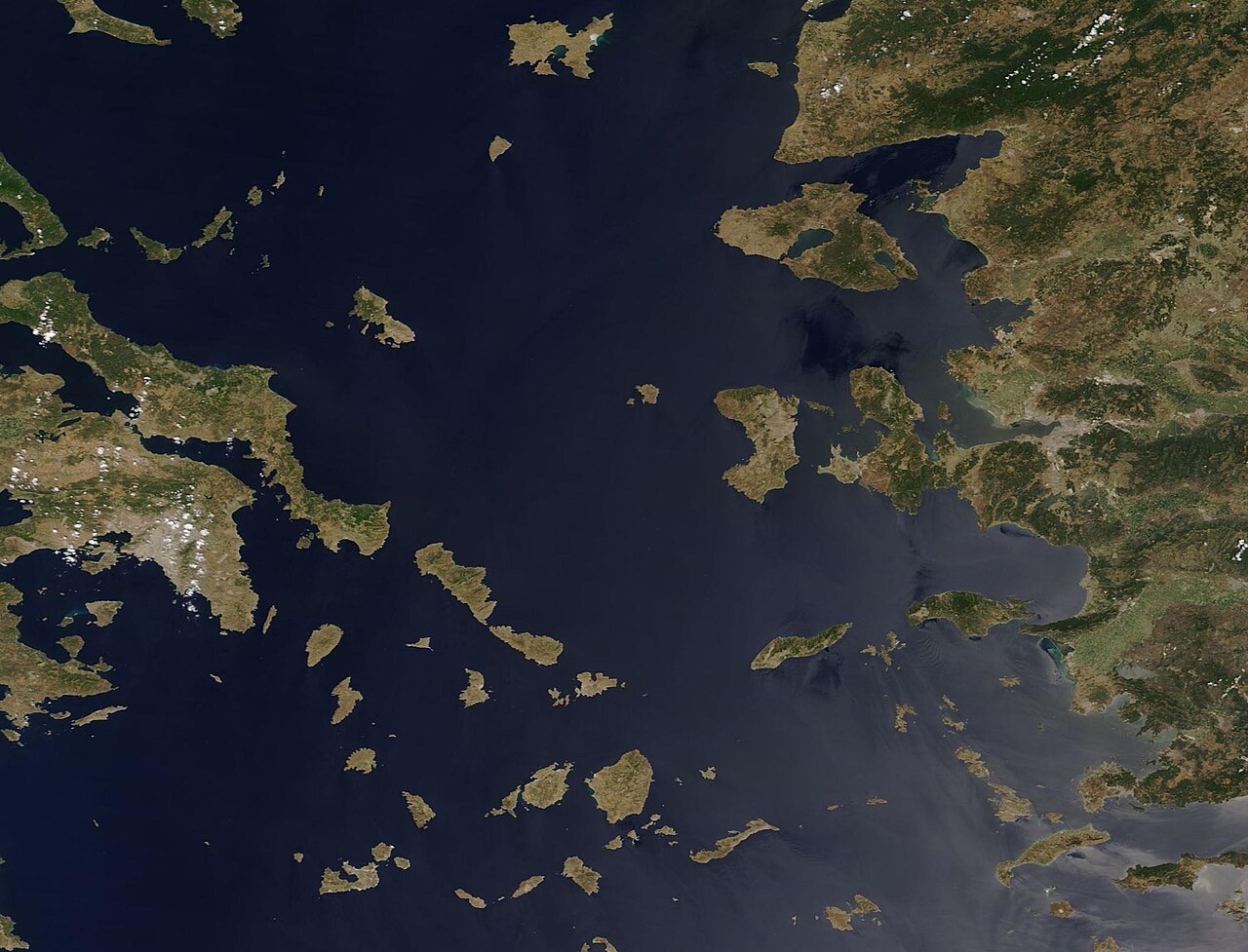 A satellite photograph of the deep blue Aegean Sea scattered with brown islands, the Greek mainland at left and the Turkish coast at right, with Lesbos lying close against Turkey near the top