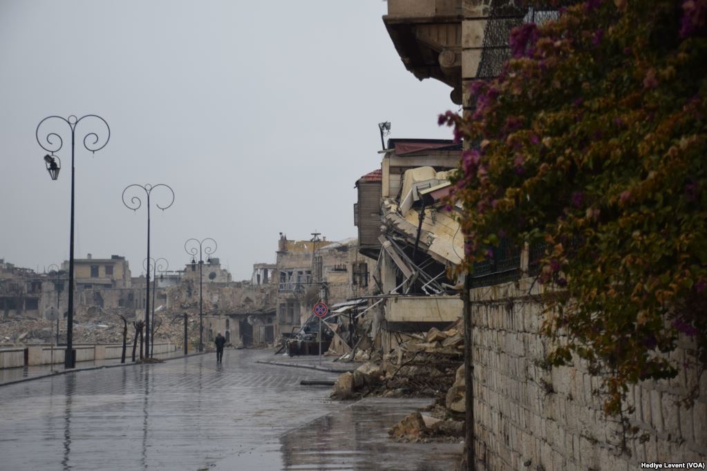A wet, empty street in Aleppo's Old City under a gray sky: whole blocks collapsed into rubble on the left, a sagging ruined building at right, and a single person walking in the distance