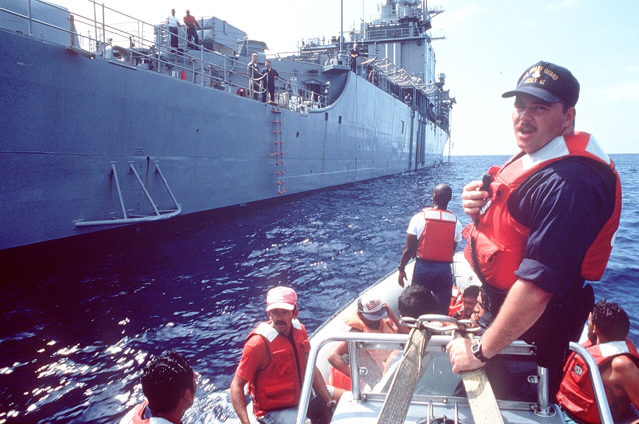 A Coast Guardsman in an orange life vest stands in a small boat crowded with rescued Cuban rafters in life jackets, alongside the gray hull of a Navy ship with a rope ladder