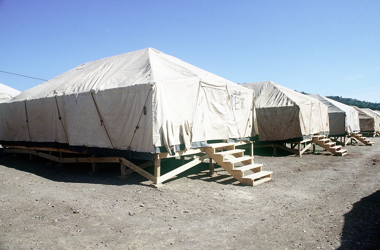 Rows of large tan strongback tents on wooden platforms with plywood steps stand on bare dirt under a clear sky, with a scrubby hillside behind