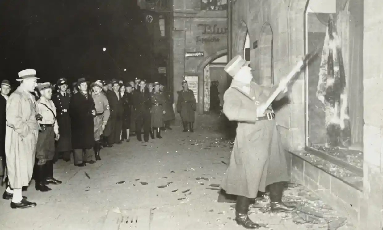 A night photograph of a man in uniform swinging a long pole at a shattered shop window while a crowd of civilians and stormtroopers stands watching on a glass-strewn street