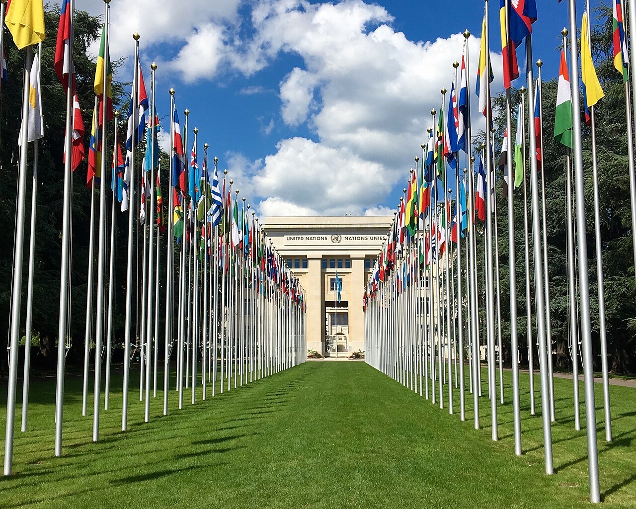 Two long rows of member-state flags on tall poles lead across a lawn to a stone building whose facade reads United Nations in English and French