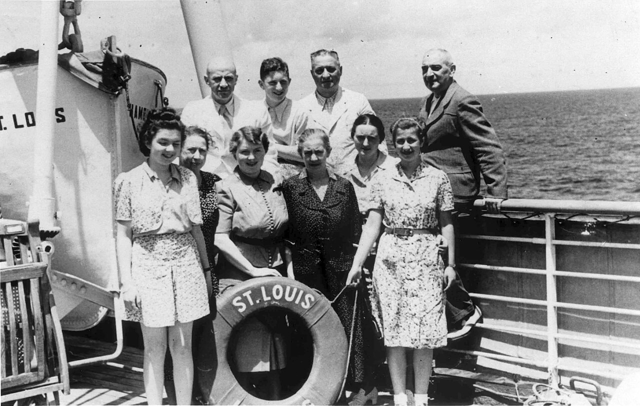 A black-and-white photograph of nine passengers posing at a ship's rail around a life ring marked St. Louis, with a lifeboat labeled St. Louis Hamburg behind them and open ocean beyond