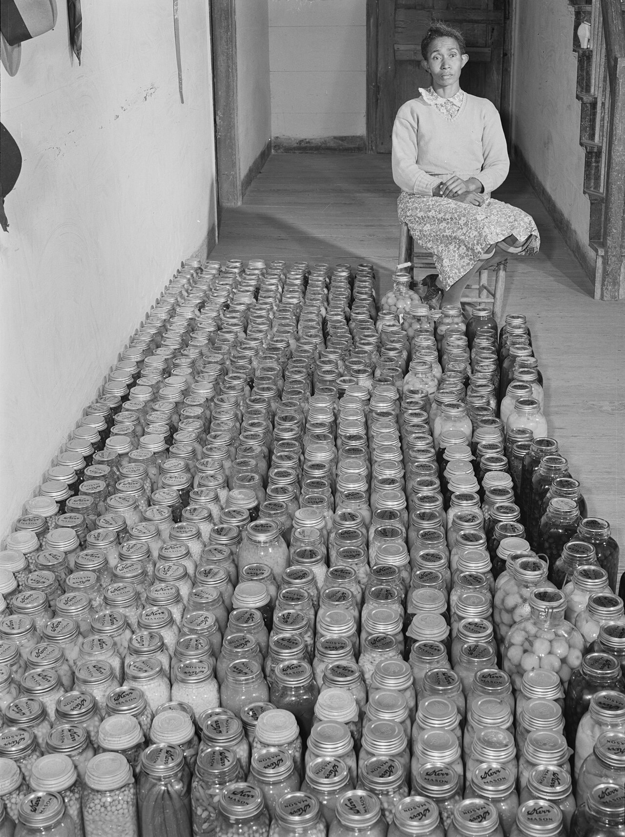 A black-and-white photograph of a farmhouse hallway whose floor is covered end to end with hundreds of Mason and Kerr canning jars, a woman seated on a chair behind them