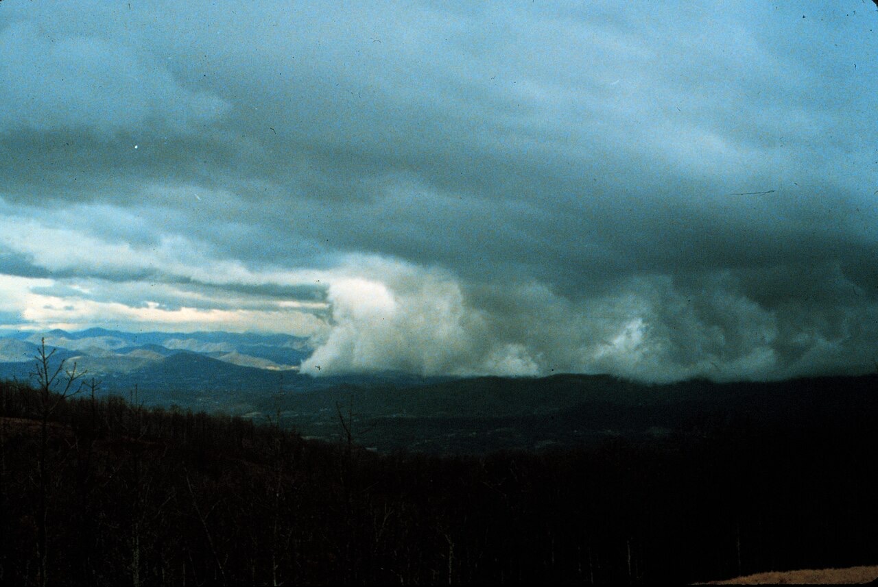 A color photograph of a dark, flat-bottomed deck of cloud advancing over wooded ridges, its pale leading edge boiling upward, with distant blue hills still in clear air at the left