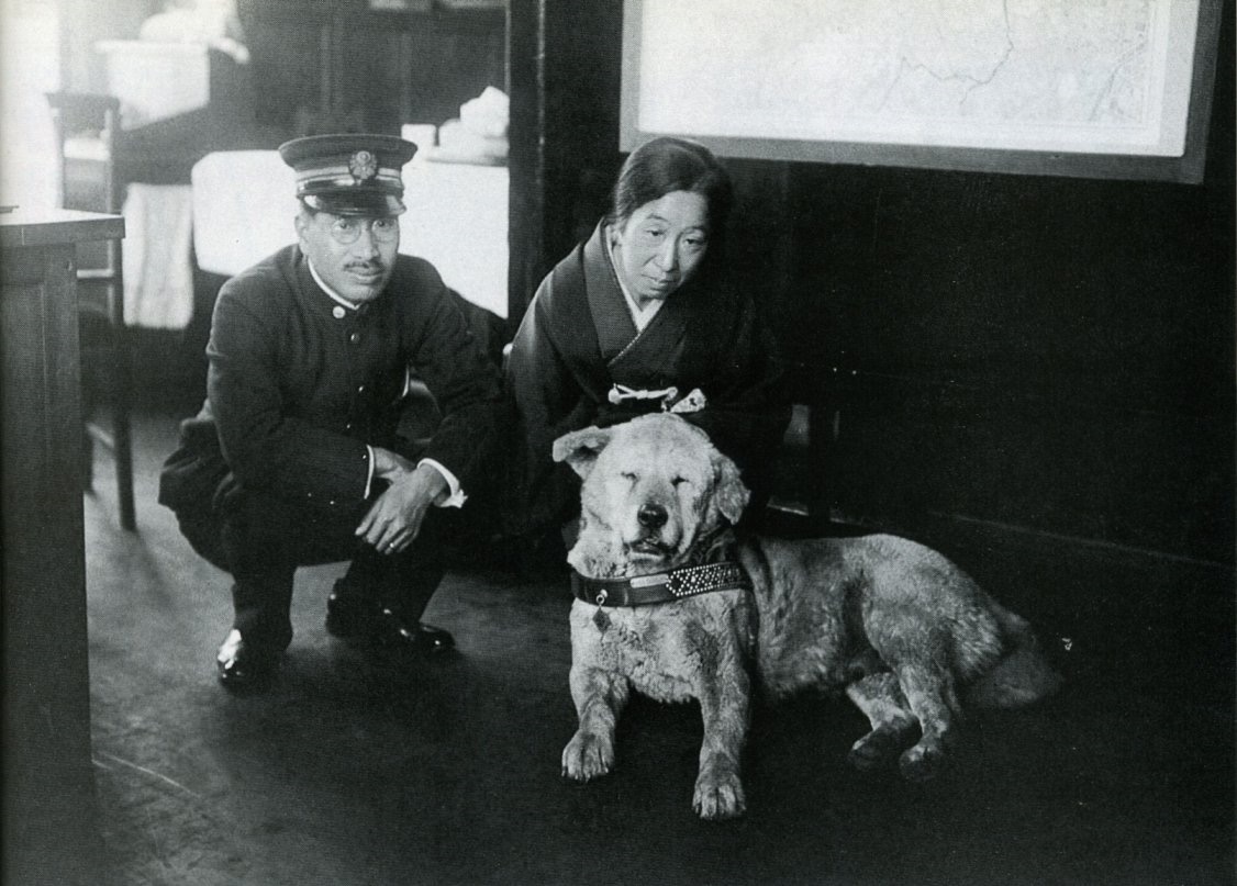 A black-and-white photograph of an old, thickset Japanese dog lying on a station floor, a uniformed stationmaster crouching at the left and a woman in a kimono resting a hand on the dog's back