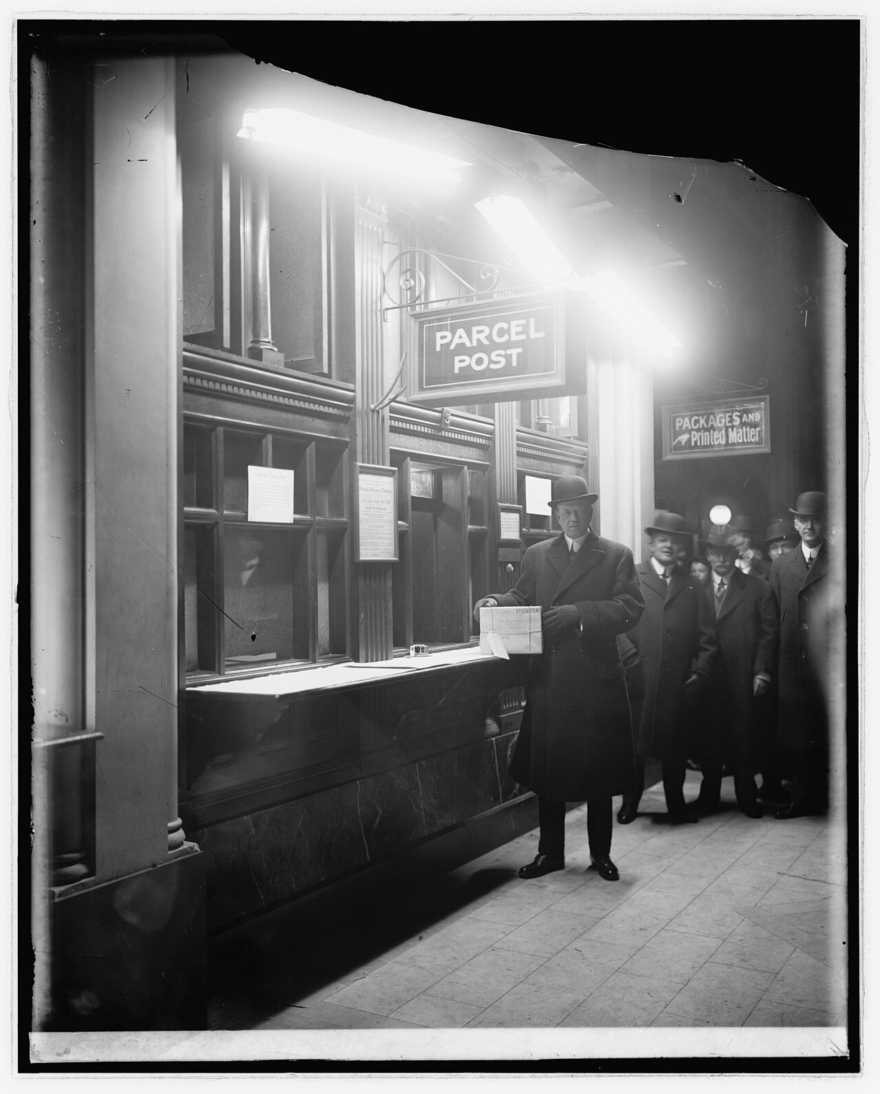 A black-and-white photograph of a post office window under a large sign reading PARCEL POST, with a man in an overcoat and bowler hat holding a wrapped, addressed package and other men watching from the right