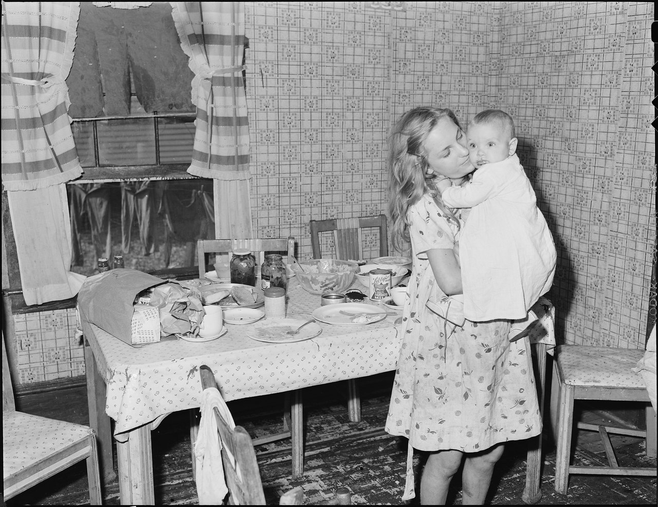 A girl in a flowered dress stands in a kitchen holding a baby against her shoulder beside a table set with dishes
