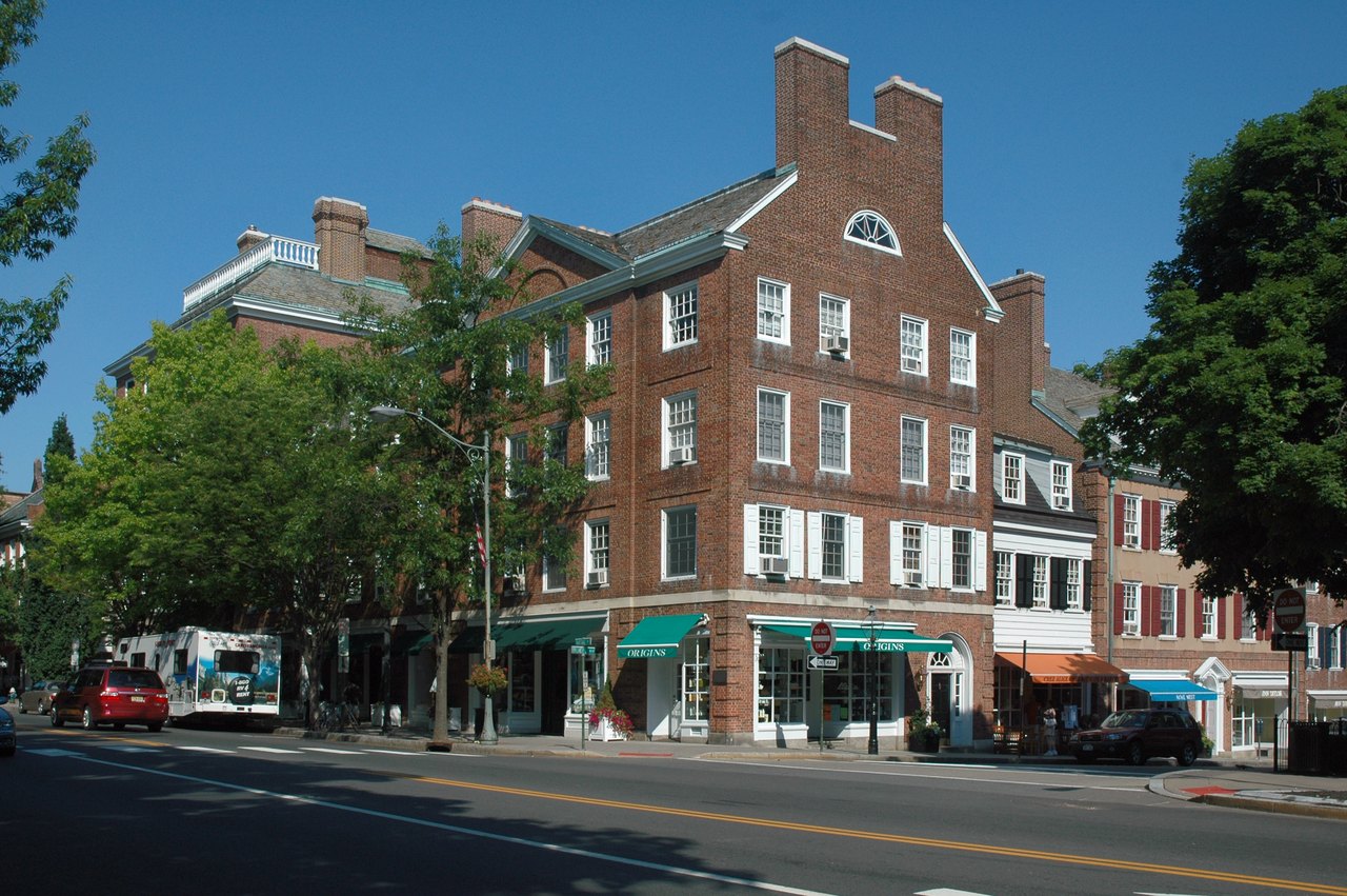 A sunny corner of a small-town main street with three- and four-story brick buildings, shop awnings, leafy trees, a bus and parked cars