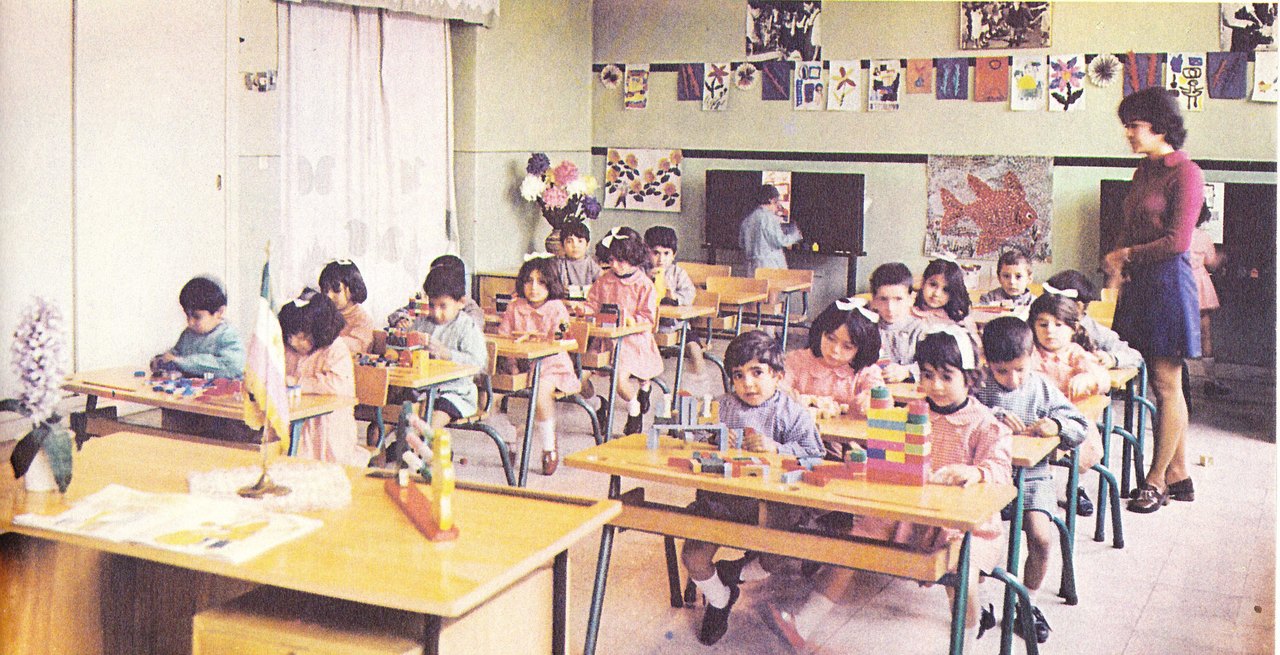 A color photograph of a preschool classroom where about twenty small children sit at low tables playing with blocks while a teacher stands at the side