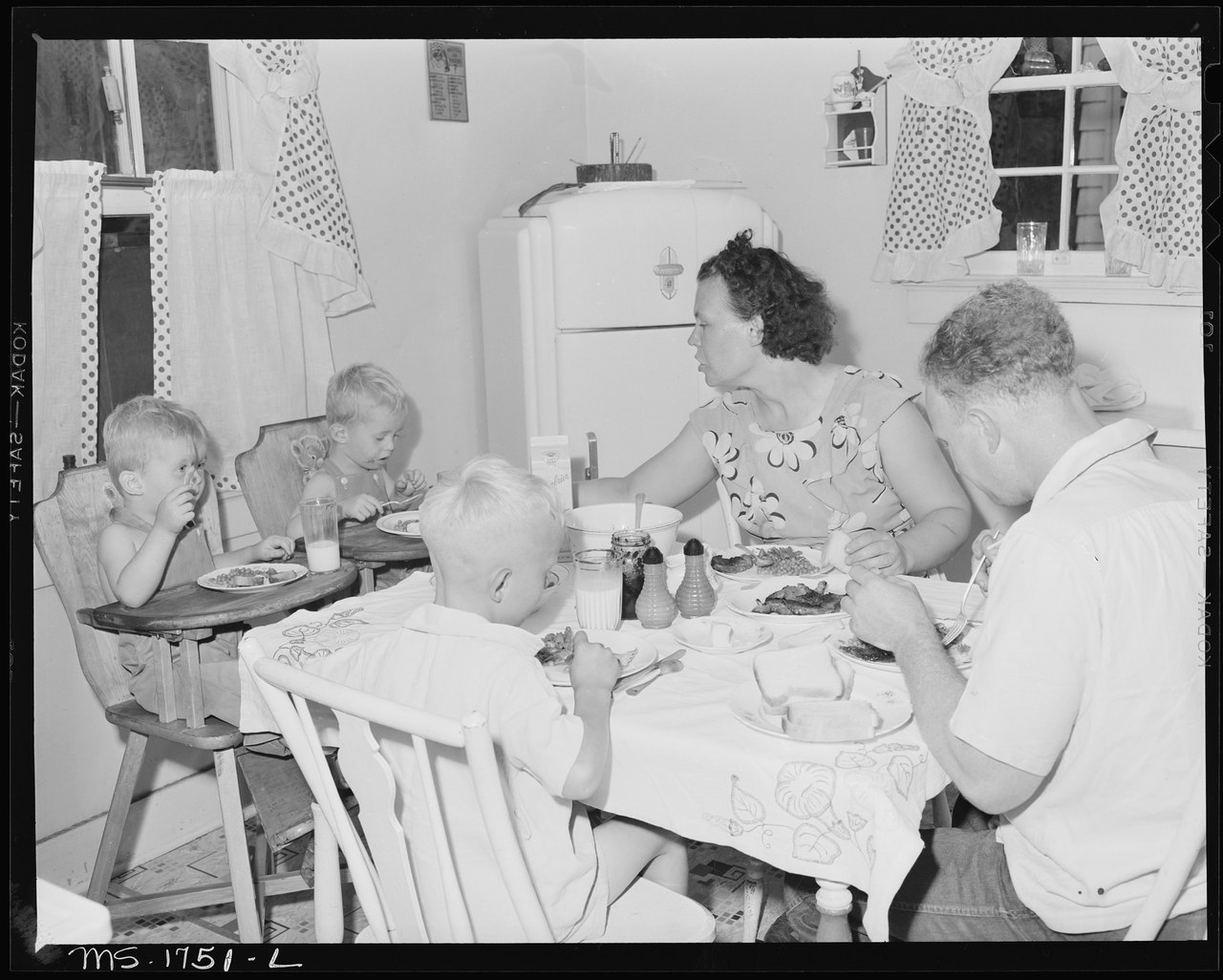 A black-and-white photograph of a family of five, including a toddler in a high chair, eating dinner around a small kitchen table in a plain, tidy kitchen