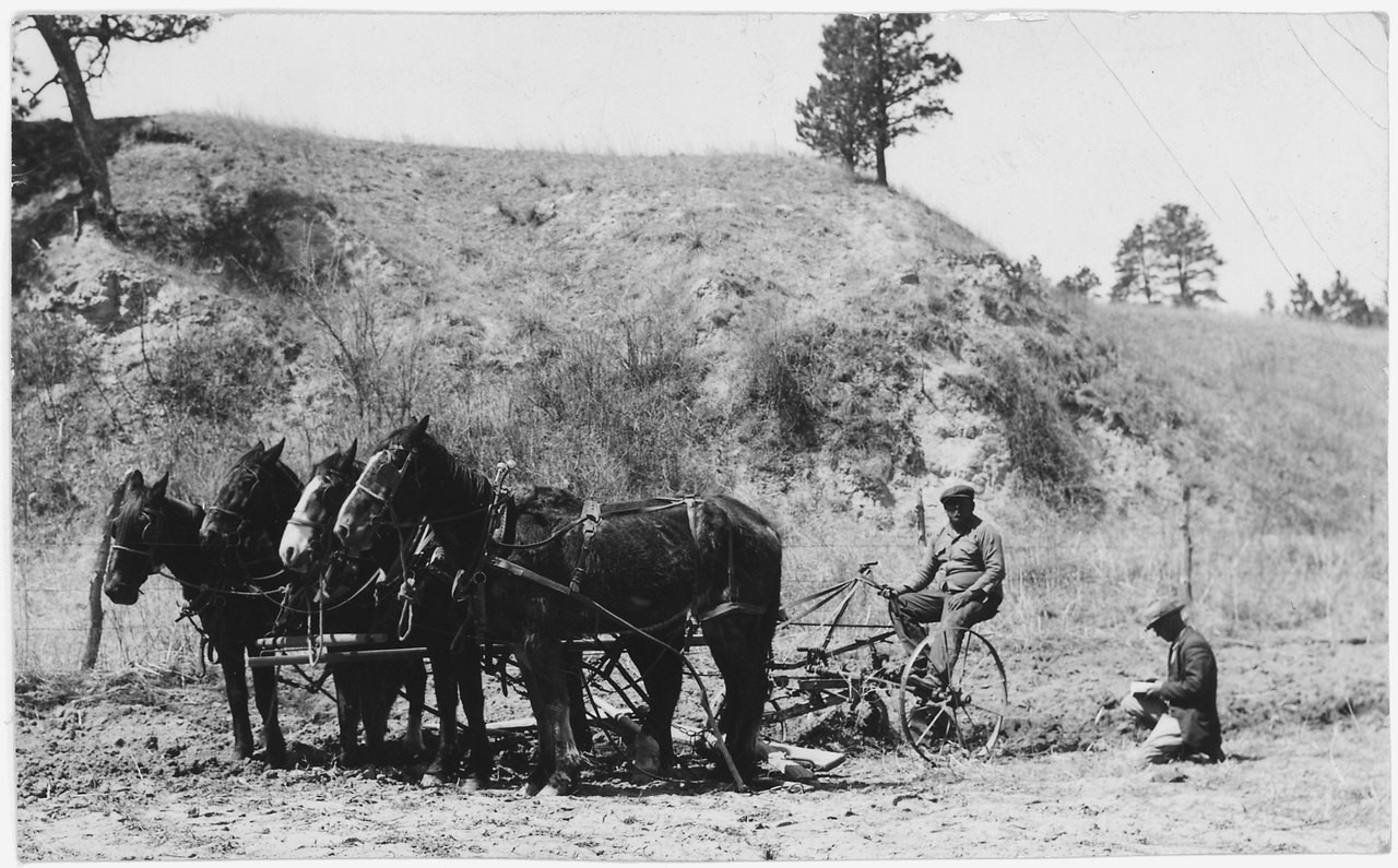 A black-and-white photograph of a farmer walking behind a plow drawn by two horses across a bare hillside field, with a second person seated at the right
