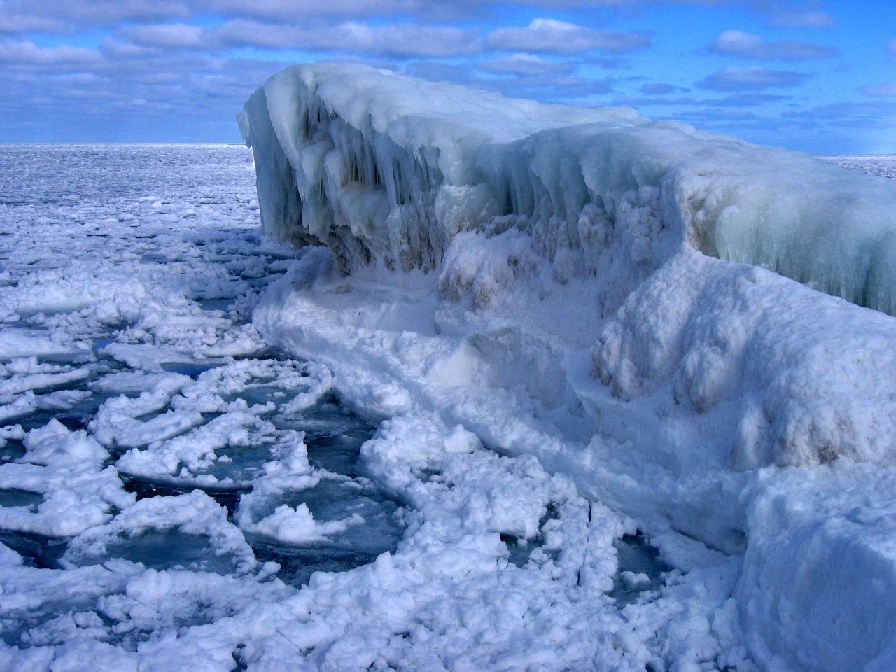 A slab of pale ice juts up at the edge of a frozen lake, with flat oval plates of pancake ice spread across the water behind it under a blue sky