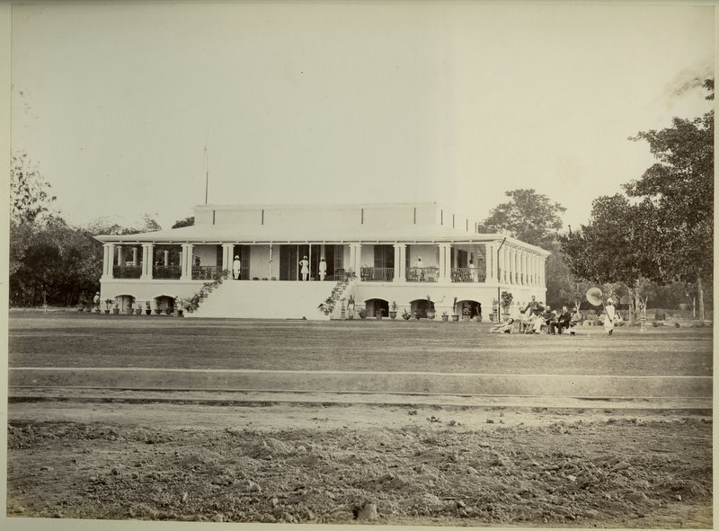 An 1870s photograph of a long, low white bungalow in India with a deep pillared veranda across its whole front, servants in white standing under the arches, and a group of people seated on the grass at one end