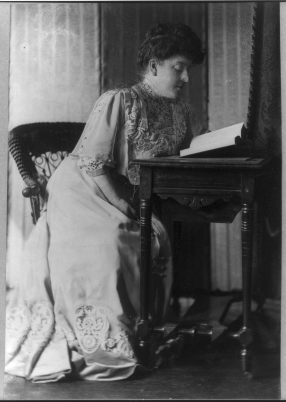 A black-and-white photograph of a woman in an embroidered light-colored gown seated on a chair beside a small table, bent forward over an open book, in profile against a curtained background