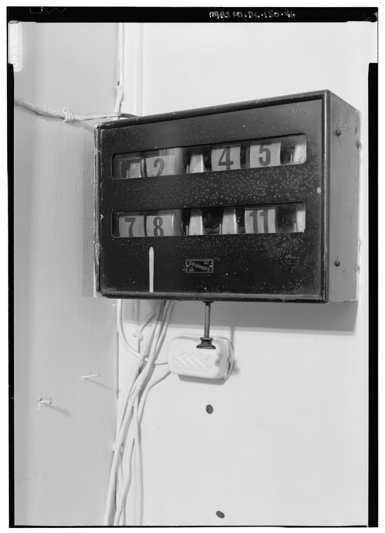 A wall-mounted metal annunciator box with two rows of small numbered windows behind glass, a maker's plate at the bottom, exposed wiring running down the wall and a push button below
