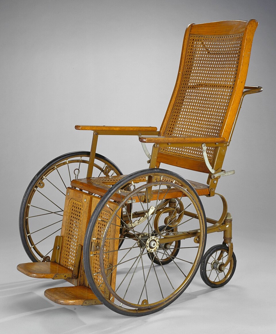 A museum photograph of a wooden wheelchair with caned back and seat, a reclining backrest, hinged wooden footrests, two large spoked wheels with solid tires and a small caster at the rear, against a plain gray background