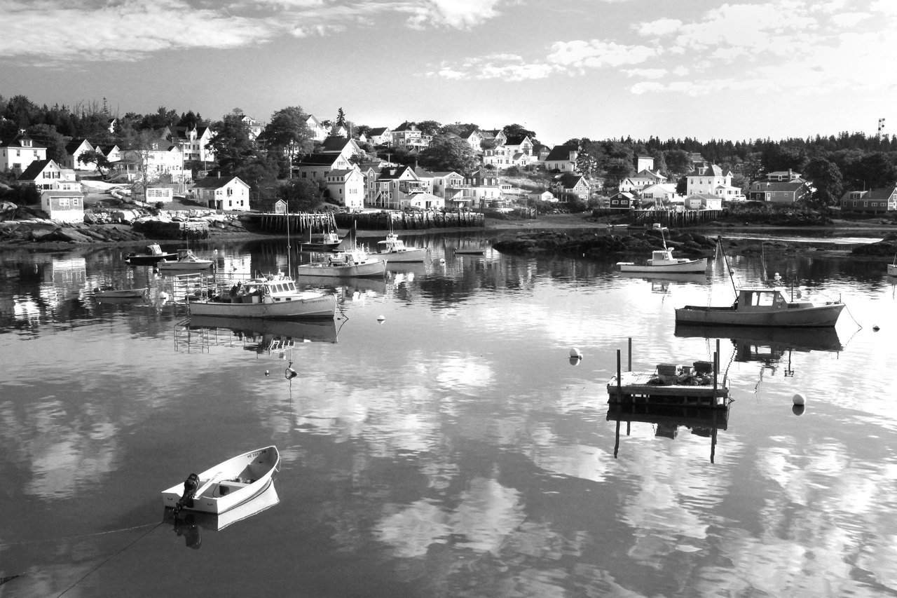 A black-and-white photograph of small boats moored on calm water in a harbor, with the houses of a small town along the far shore under a cloudy sky