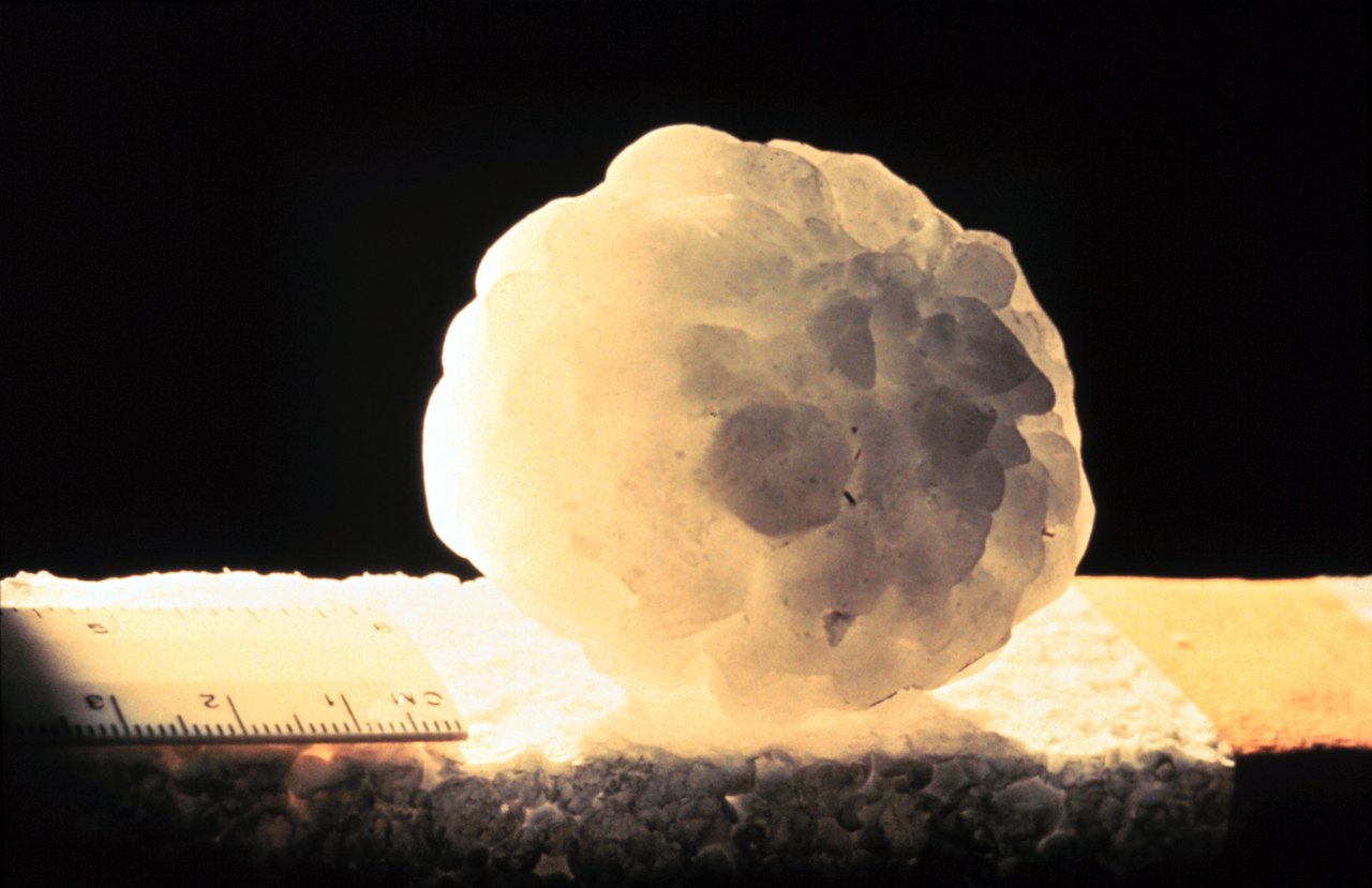 A lumpy white hailstone made of many smaller stones fused together sits on a dark surface beneath a ruler, with a label reading Ada, Oklahoma, 29 April 1978