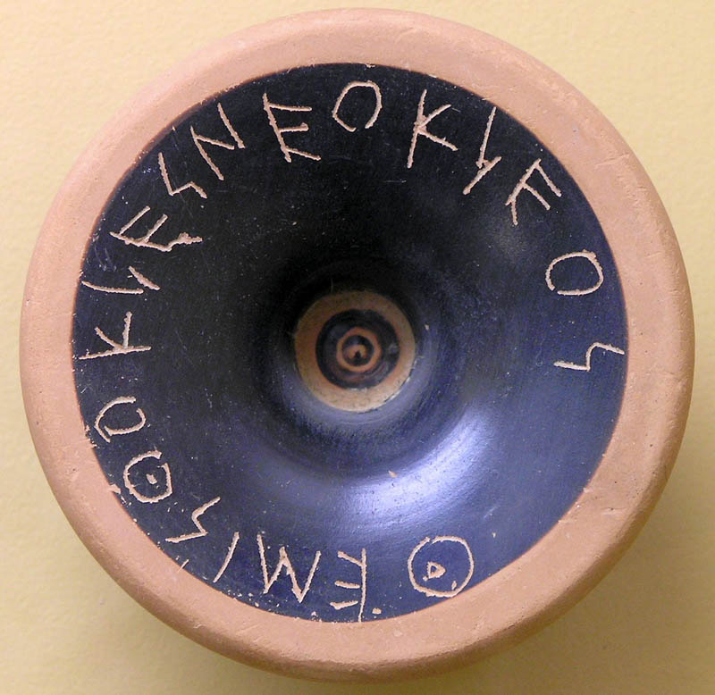 The round base of an ancient black-glazed pottery vessel with Greek letters scratched around its rim, spelling the name Themistocles