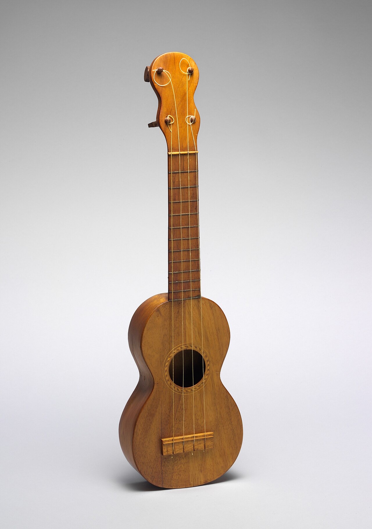A small four-stringed wooden ukulele with a curved figure-eight body, photographed upright against a plain gray background