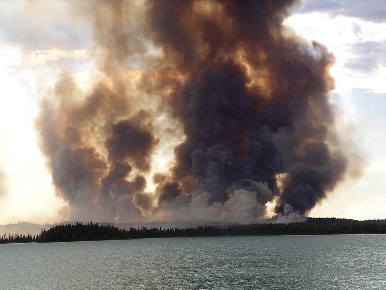 Towering brown and gray smoke columns from a forest fire rising over a dark line of spruce trees, seen across the still water of a lake