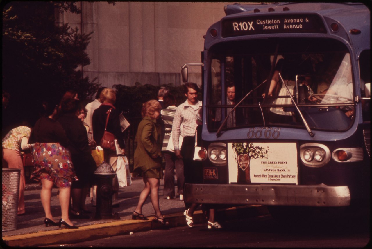 A line of people in 1970s clothes step up into a city bus at a curb beside a stone building; a woman in a flowered dress is at the front of the line