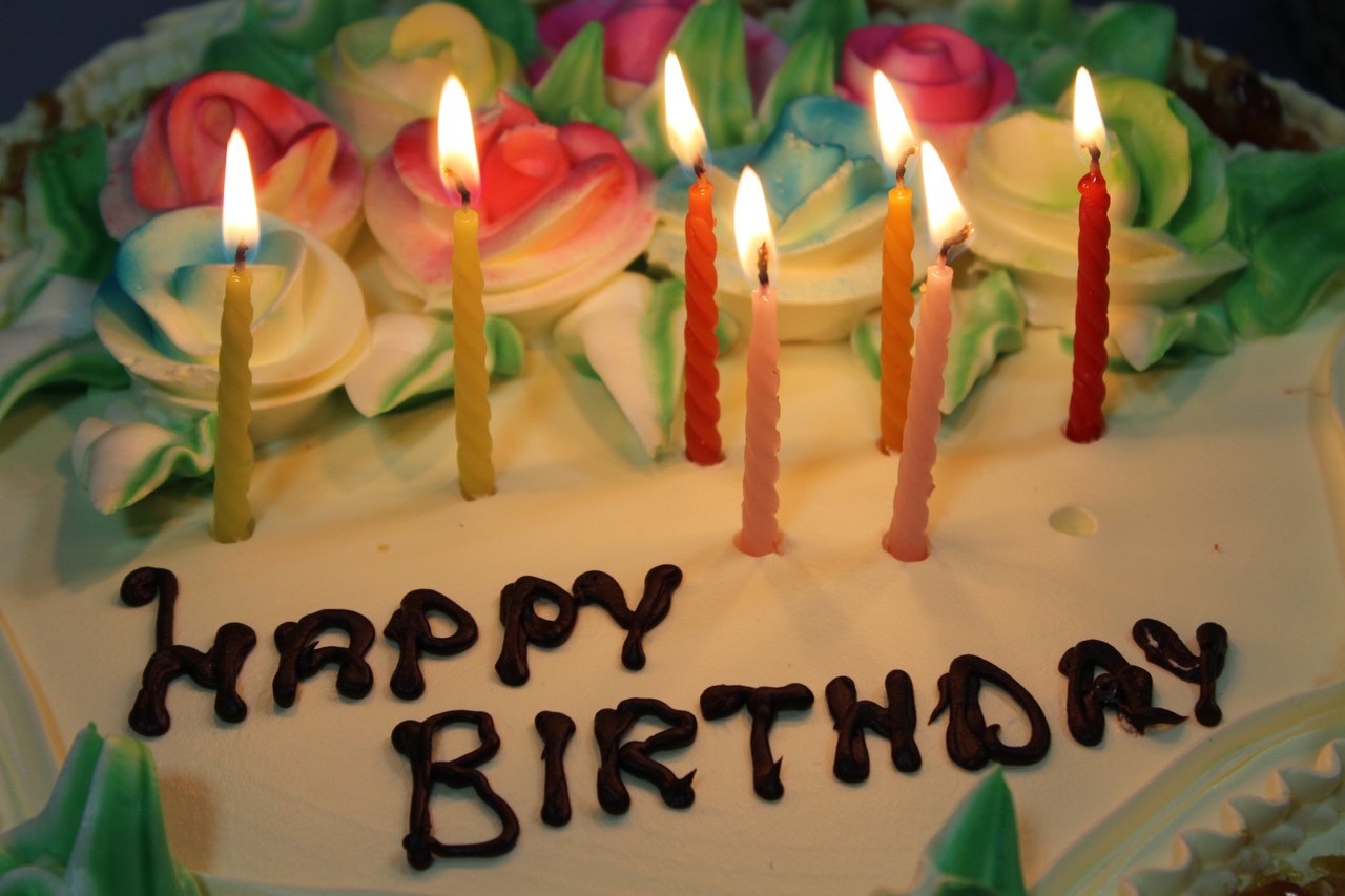 Close-up of a white frosted cake decorated with pink and green icing flowers and the words HAPPY BIRTHDAY, with seven thin lit candles standing in it