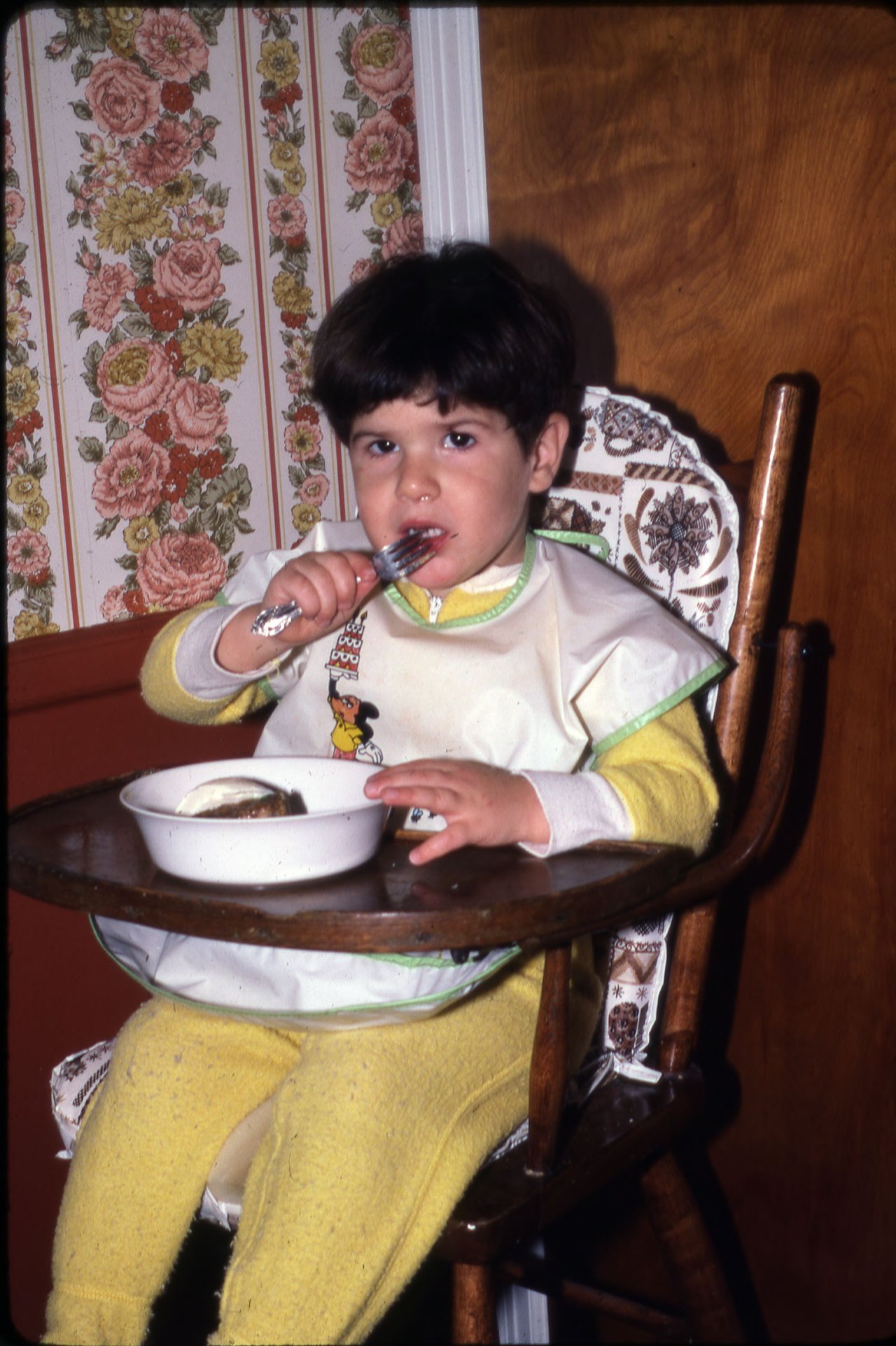 A small dark-haired child in yellow pajamas and a bib sits in a wooden high chair, holding a fork to his mouth over a bowl, with flowered wallpaper behind
