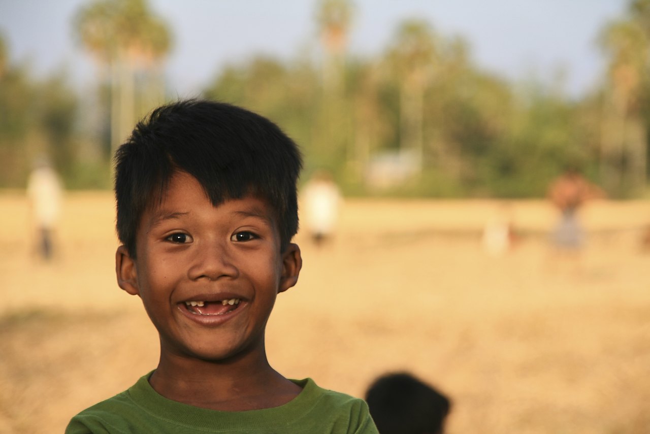A laughing boy in a green shirt, photographed outdoors in a sunny field, shows a wide smile with a gap where his two upper front teeth are missing