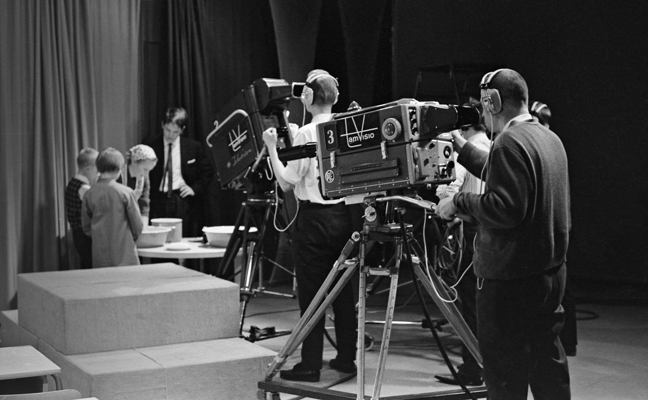 Black-and-white view of a television studio: two camera operators in headphones stand behind large boxy cameras on wheeled stands, aimed at a lit set where three small children and a man in a suit stand around a low table with bowls