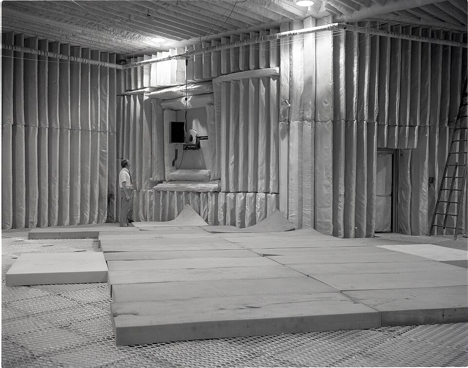 A black-and-white photograph of a large test chamber whose walls and ceiling are covered in deep sound-absorbing baffles, with a man standing on foam panels laid over a mesh floor