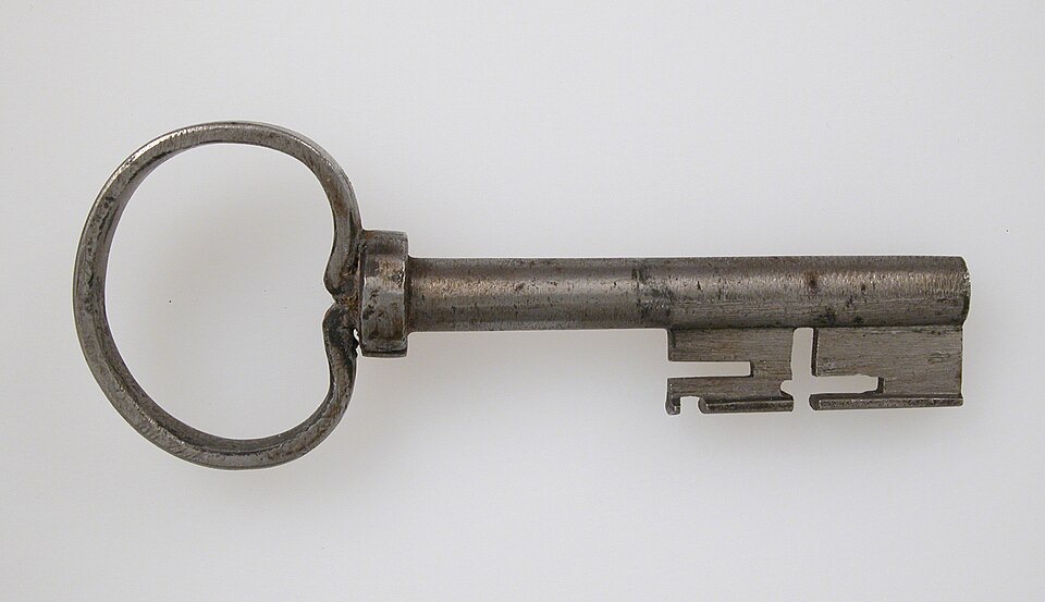 A hand-forged iron key with a large oval bow, a stout round shaft, and a squared bit with deep slots, photographed on a plain pale background