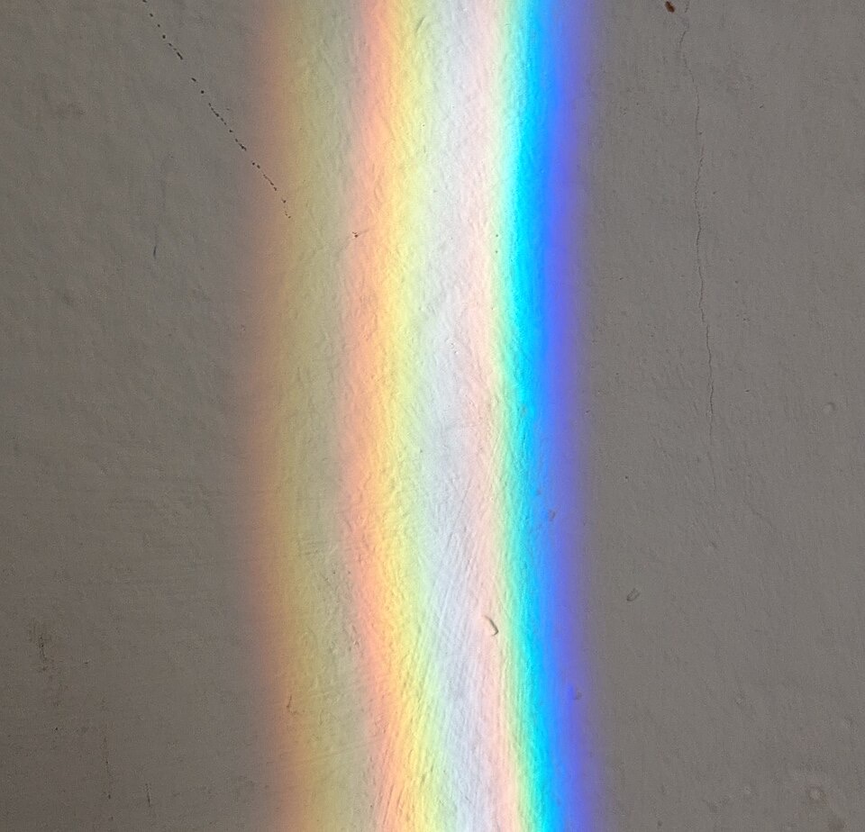 A broad band of rainbow colors, red through violet, cast by sunlight onto a rough white plaster wall with fine cracks