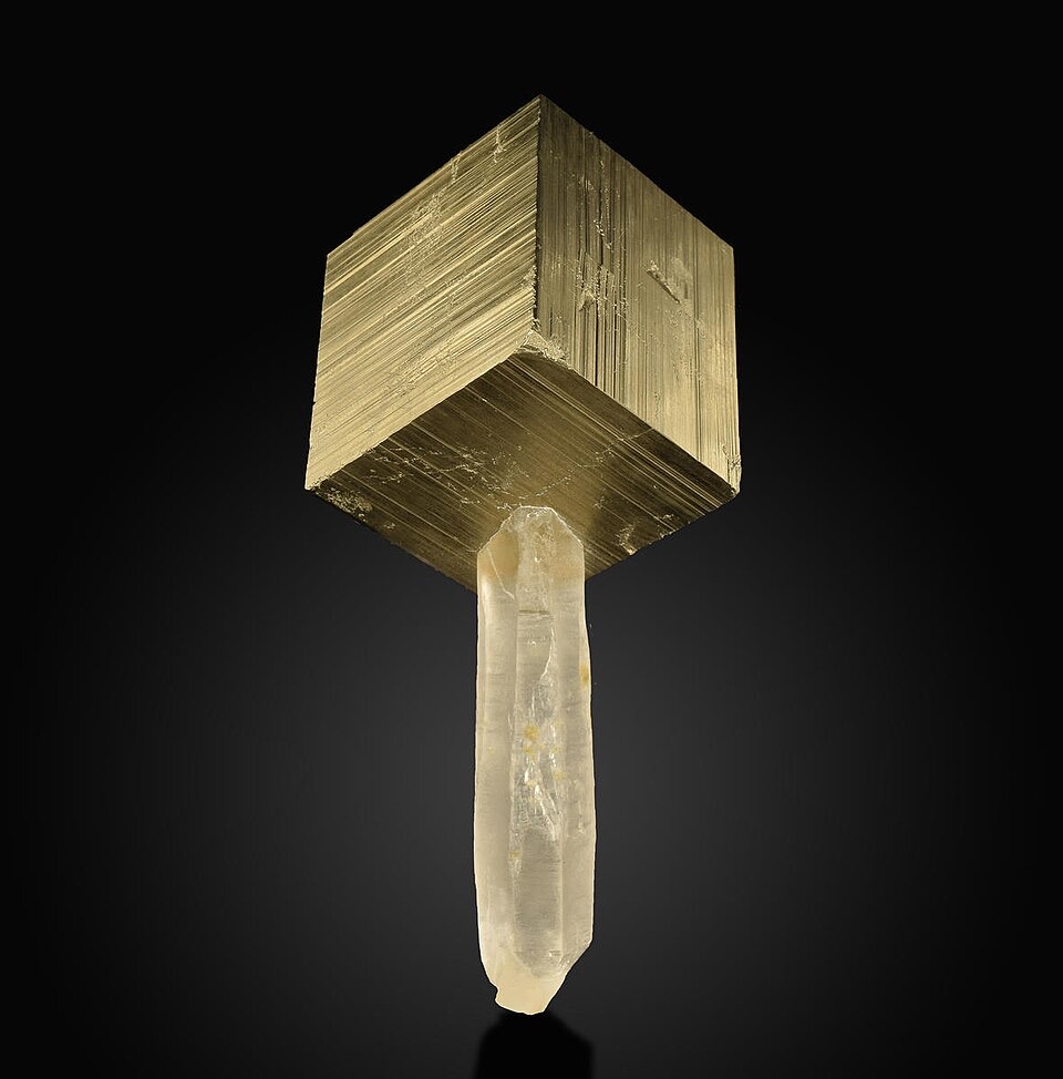 A brassy metallic cube with finely striated faces perched on a translucent white quartz crystal, photographed against a black background