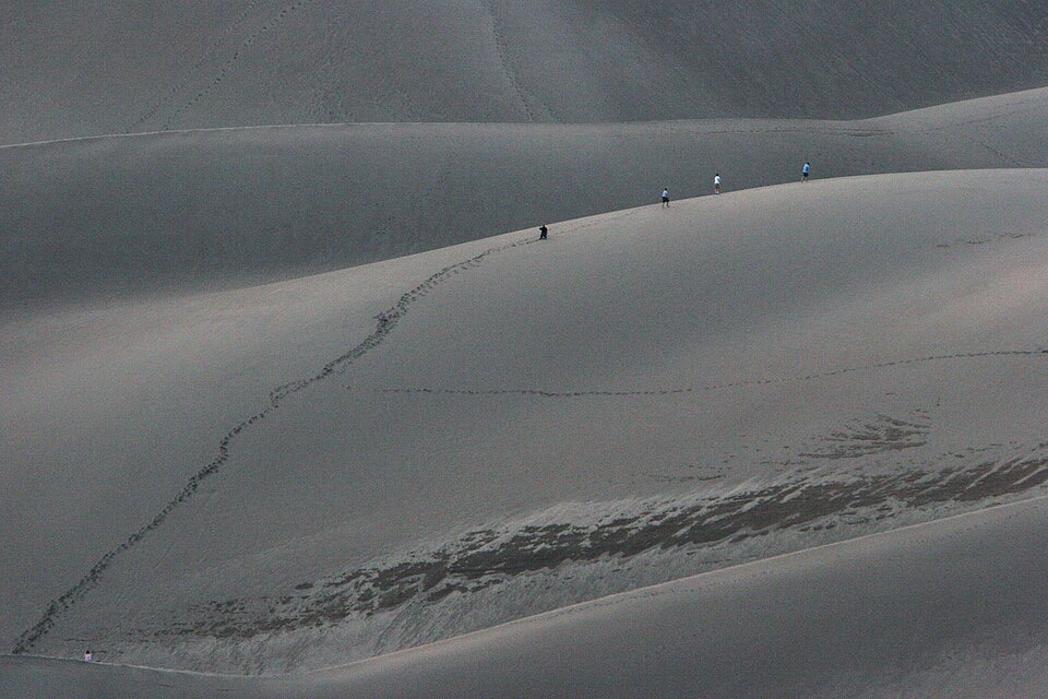 Vast smooth sand dunes filling the whole frame, with a winding trail of footprints and five hikers so distant they are tiny dots