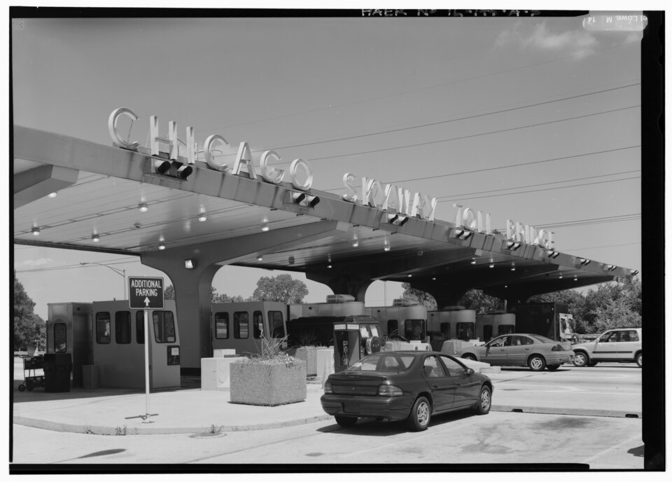 A black-and-white photograph of a highway toll plaza: a row of small glass booths under a wide winged canopy lettered CHICAGO SKYWAY TOLL BRIDGE, with a car passing through