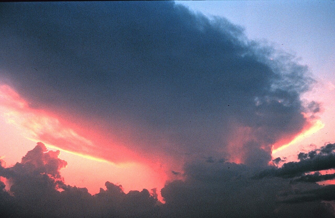 A towering storm cloud at sunset, its dark flat top spreading sideways like an anvil while the clouds below glow pink and orange