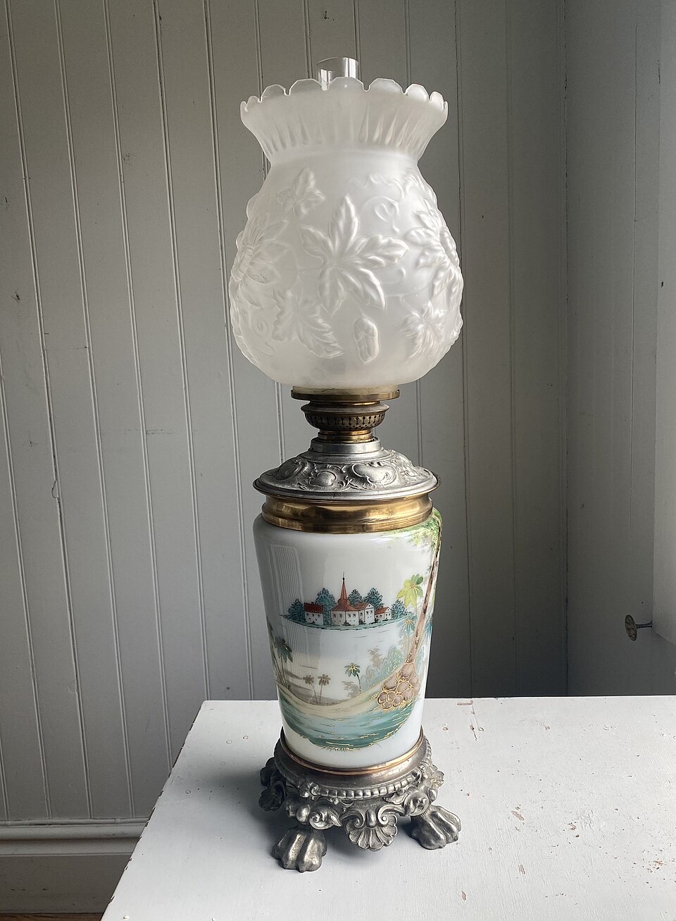 An ornate parlor kerosene lamp with a clawed metal foot, a painted ceramic body, a brass burner and a tall frosted glass shade, standing on a white table