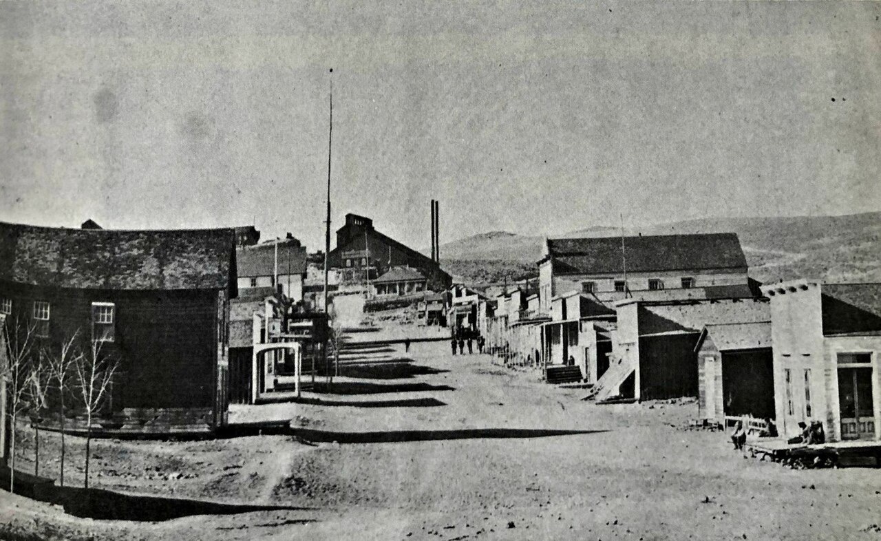A grainy black-and-white photograph of a wide dirt main street lined with false-front wooden and brick buildings, with hills and a mill building beyond