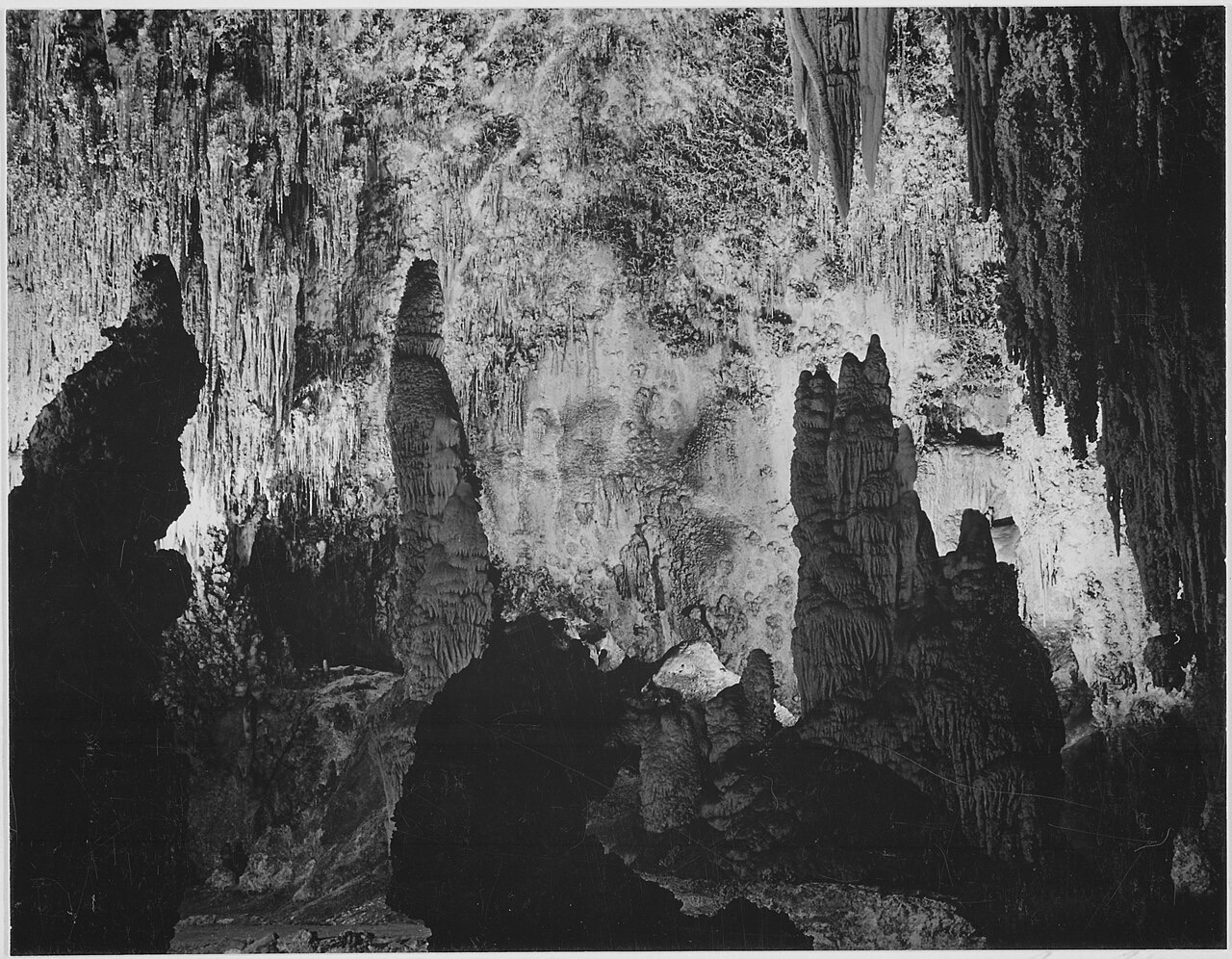 A black-and-white photograph of a cave chamber crowded with thin stalactites hanging from the ceiling and massive stalagmites rising from the floor