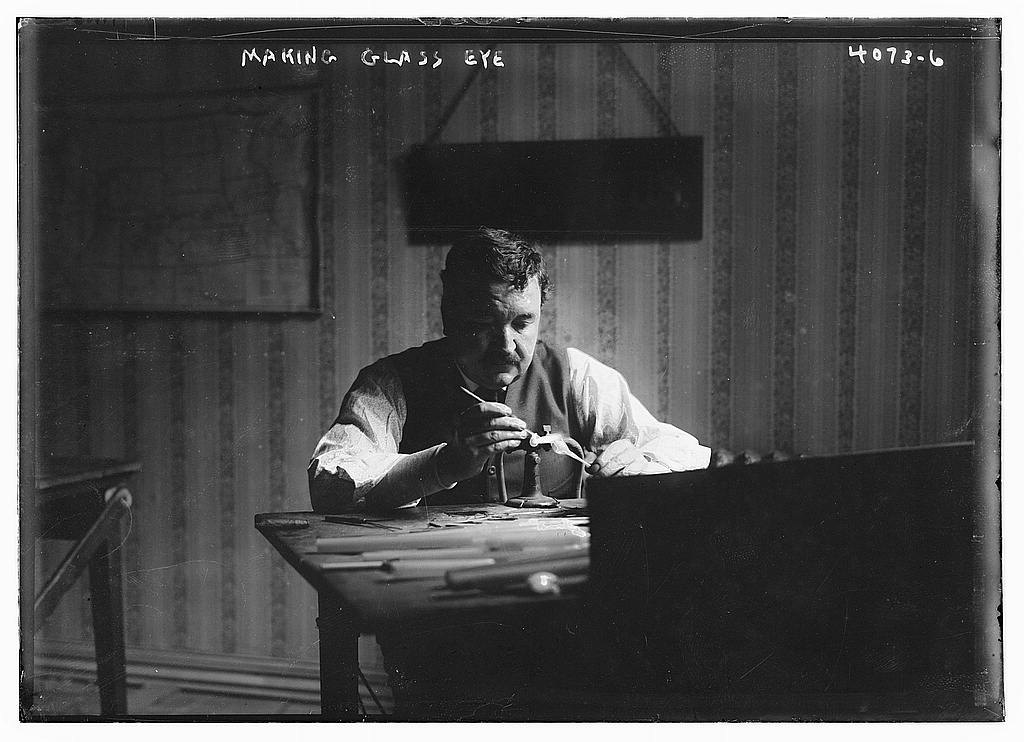 A black-and-white photograph from about 1915 of a man in shirtsleeves seated at a workbench, bending close over a small flame and tool as he shapes a glass eye
