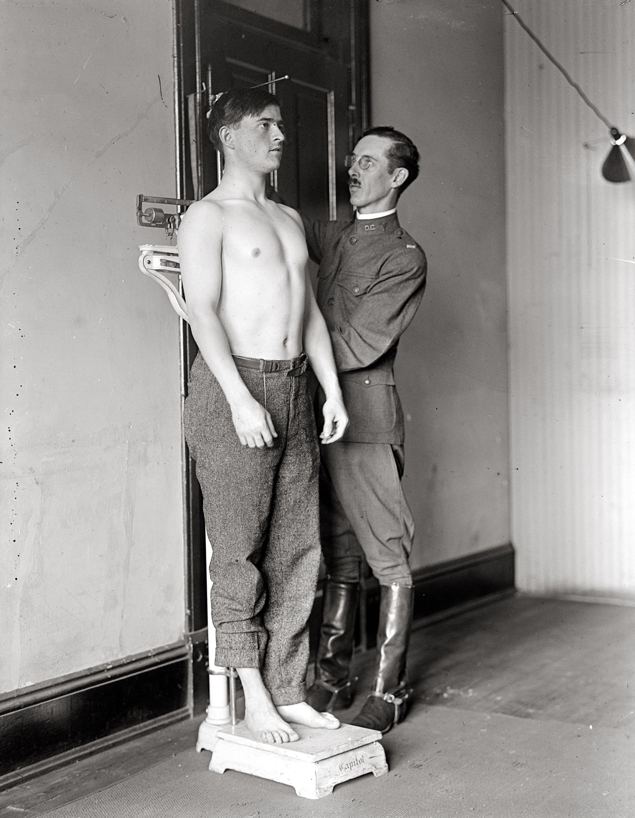 A black-and-white 1917 photograph of a shirtless young man standing on a scale against a wall while a uniformed doctor lowers a measuring bar onto his head