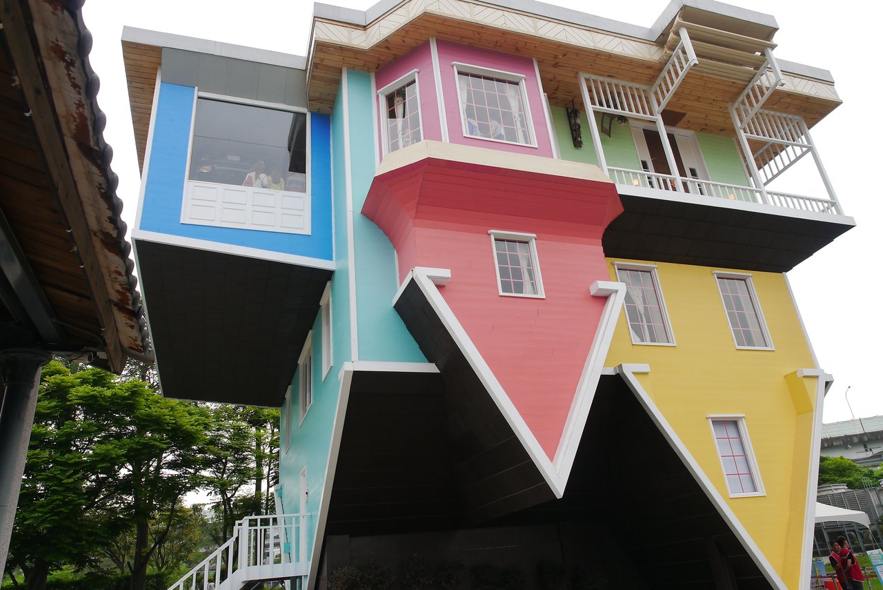 A brightly painted two-story house standing on its pointed roof, with its walls, windows and balcony all upside-down against a blue sky and trees
