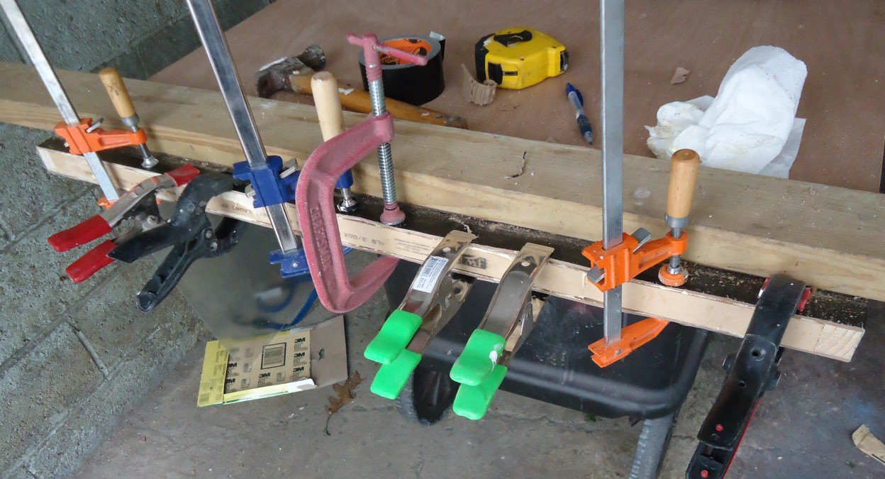A row of metal and plastic clamps in orange, green and red squeezing a long strip of wood along the bottom edge of a door lying on a workbench