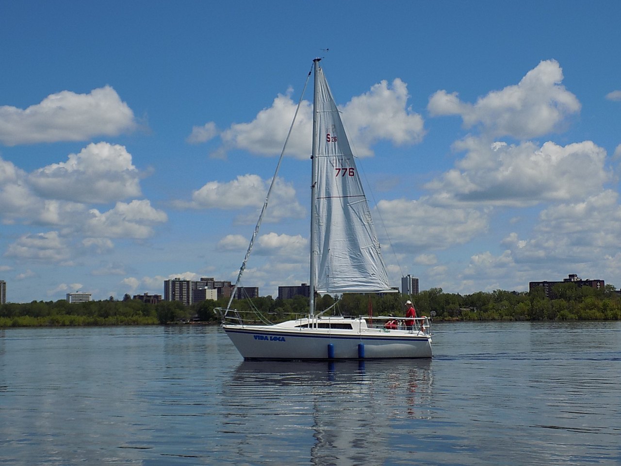 A small white sloop under sail on flat water, its tall mast carrying a large mainsail behind and a smaller triangular jib in front