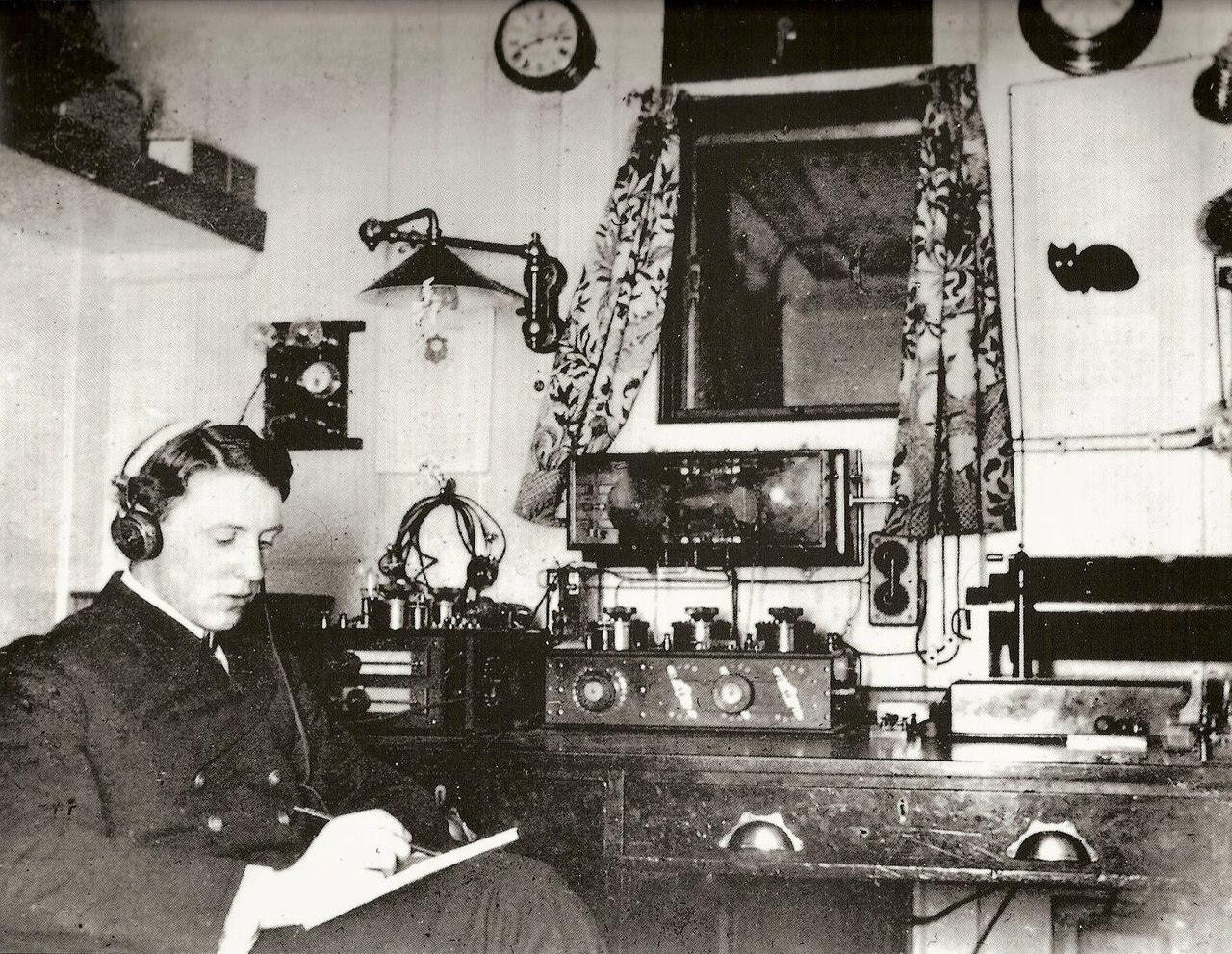 A ship's radio room: an operator in headphones sits writing at a bench of dials, switches and instruments, a clock on the wall above him