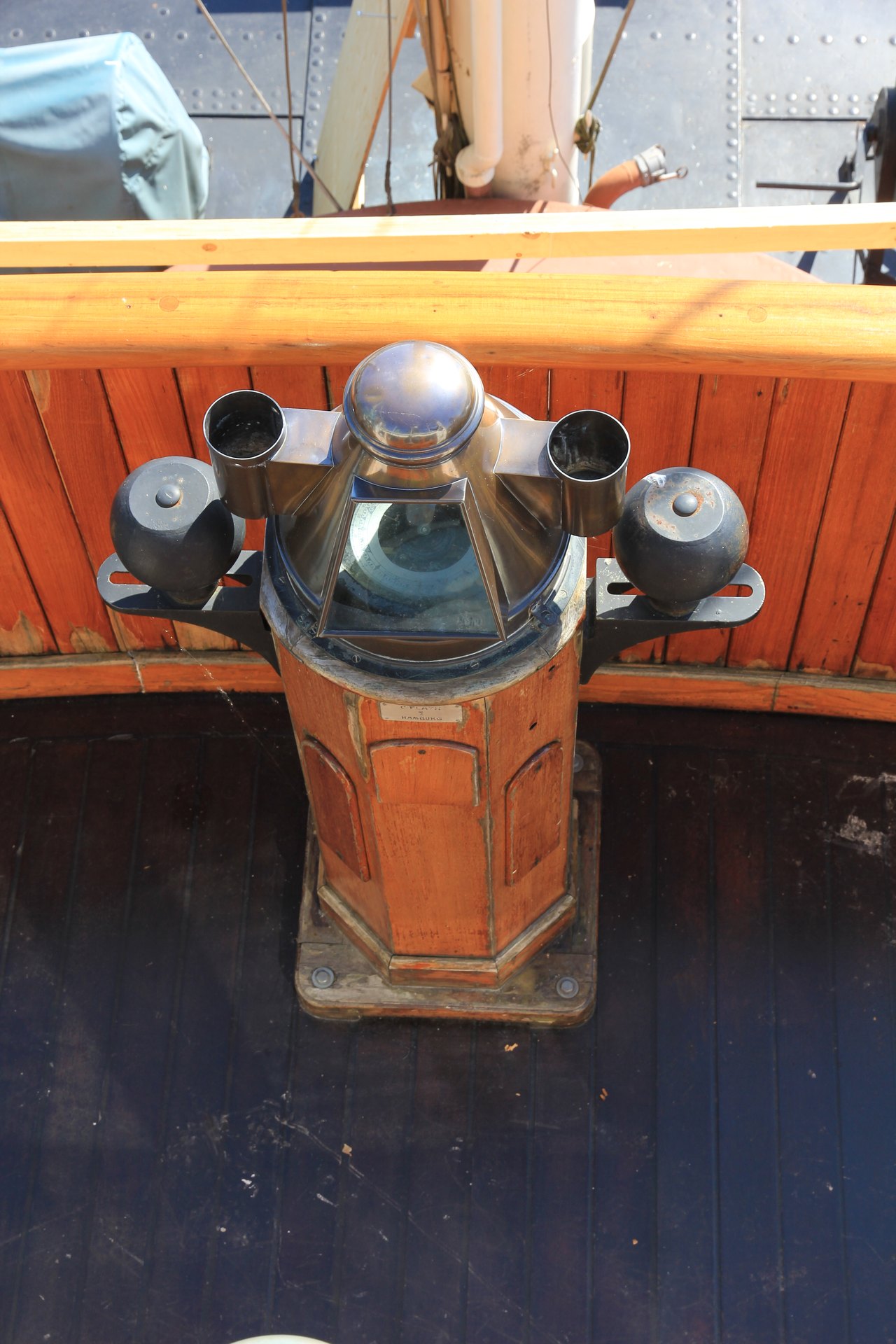 A ship's compass mounted in a wooden binnacle on a bridge, its glass dome flanked by two dark metal spheres