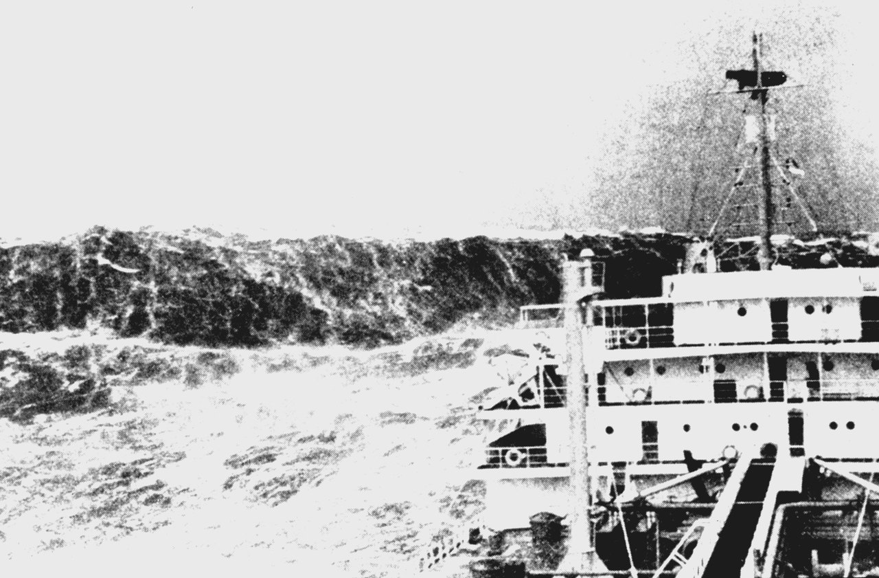 A very large wave rearing above a ship's rail and superstructure, its crest already breaking into spray as it reaches the deck