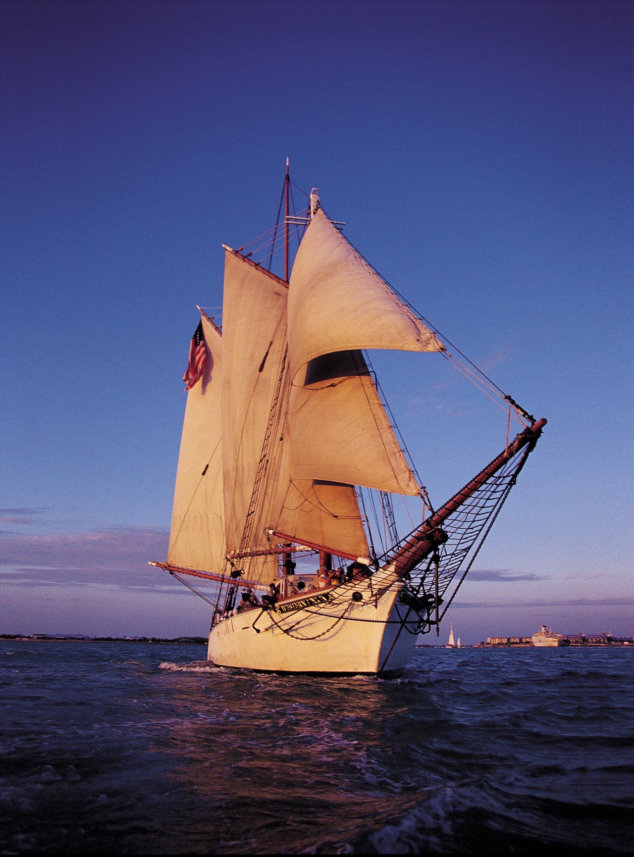 A two-masted schooner under full sail in low evening light, heeling slightly, with land just visible on the horizon behind her